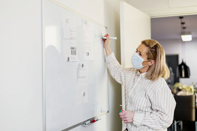 Woman wearing protective face mask writing on whiteboard