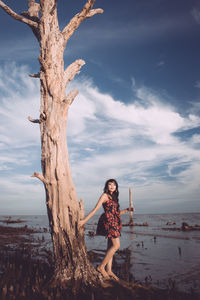 Portrait of young woman standing by tree trunk near sea against sky