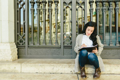 Full length of woman writing in book while sitting on footpath