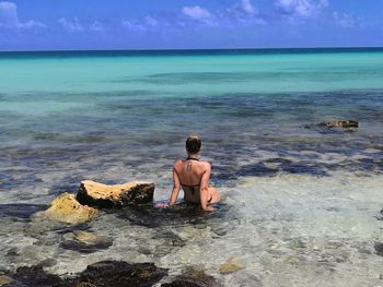 Rear view of woman in bikini sitting at beach against blue sky