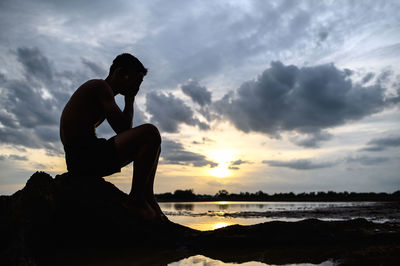 Silhouette boy sitting on rock against sky during sunset