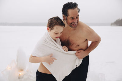 Cheerful shirtless mature man standing with son wearing towel at frozen lake