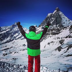 Rear view of man with arms raised standing against snowcapped mountains