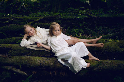 Two girls sisters in white dresses lie on logs in the moss in the forest