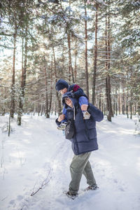 Happy single dad carries his little son on shoulders so that he can reach the branch and shake snow