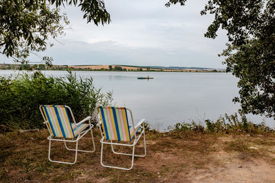 Chairs by plants on lake against sky