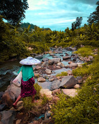 Rear view of woman standing on rock by river against sky