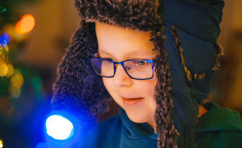 A little boy in a blue earflap hat and glasses looks at a glowing christmas tree