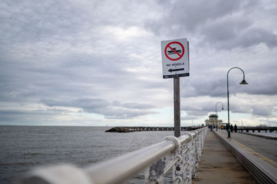 Road sign by railing by sea against sky