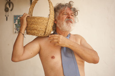 Shirtless man holding basket while standing against wall