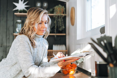 Young woman with blonde curly hair in grey sweater reading book on kitchen at house