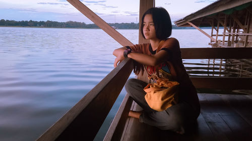 Thoughtful young woman sitting on stilt house over lake during sunset