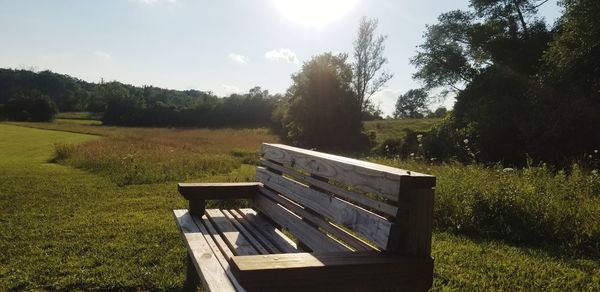 Bench on field by trees against sky