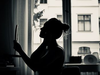 Young woman looking in mirror against window at home