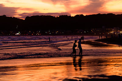 Silhouette people walking on illuminated beach against sky during sunset