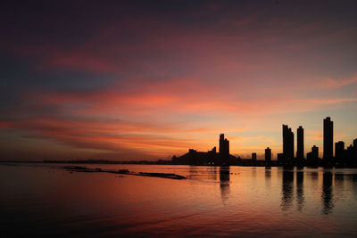 Silhouette buildings by river against sky during sunset