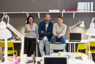 Portrait of confident multi-ethnic business colleagues sitting on desk against wall at creative office