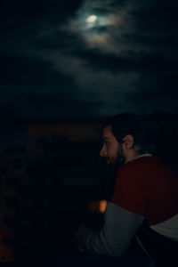 Side view of young man sitting against sky at night