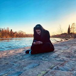 Woman using smart phone while sitting against lake during sunset