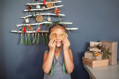 Smiling girl holding orange slice over eyes against christmas tree hanging on wall