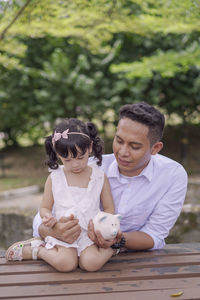 Smiling father and daughter holding piggy bank while sitting outdoors