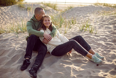 Husband and wife in hoodies and sneakers sitting on a sandy beach by the sea in autumn