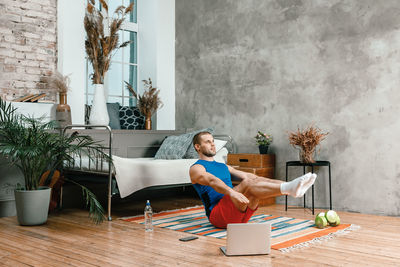 The athlete makes the press, lifting his legs up, watches a movie and social networks on laptop