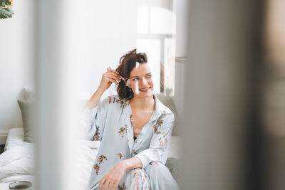 Young brunette woman in blue pajamas doing face massage with stone roller sitting on bed at home