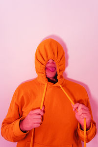 Woman wearing orange hood standing against pink wall