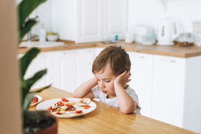 Cute toddler boy sitting with puncakes with berries in kitchen home. child doesn't want to eat