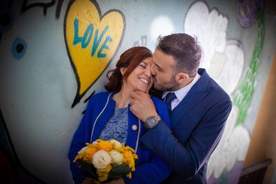 Man kissing smiling woman holding bouquet by wall