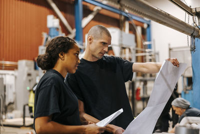 Mature man holding blueprint while standing next to female colleague in warehouse