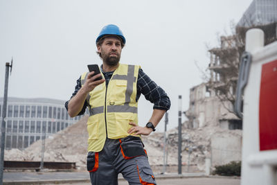 Worker holding smart phone with hand on hip in front of construction site