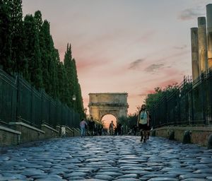 People walking on cobblestone street against sky during sunset