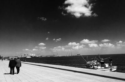 Rear view of men walking on footpath by sea against sky