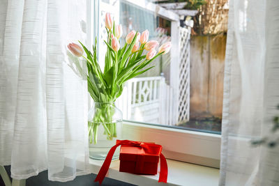 Bouquet of spring tulips in a vase on the windowsill by the window and next to it is a red gift box