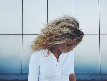 Woman with windswept curly blond hair standing against wall