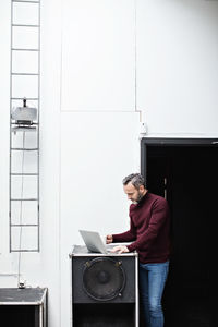 Mature businessman having coffee and using laptop while standing by wall at creative office