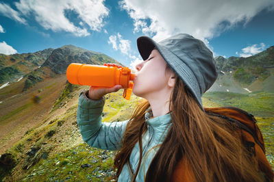 Young sports girl stands against the backdrop of a stunning mountain view and drinks clean water