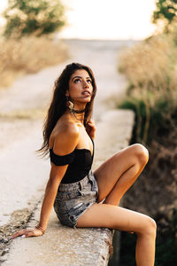 Side view of carefree barefoot young hispanic female in black top and gray denim shorts sitting on stone bridge in summer day in countryside