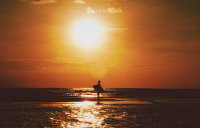 Silhouette woman with surfboard standing on shore at beach against sky during sunset