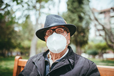 Aged man wearing protective mask with hat looking at camera while sitting on wooden bench in park on blurred background