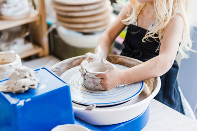 Crop unrecognizable little girl in black apron standing near pottery wheel while shaping clay pot in light workshop