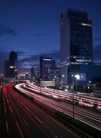 Light trails on road by buildings against sky at night