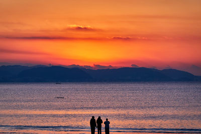 Silhouette people by sea against orange sky during sunrise