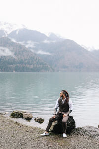 A tall brutal man hipster groom in a wedding suit stands by the misty mountains and lake in nature