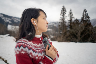 Young woman looking away while listening music over mobile phone during winter against sky