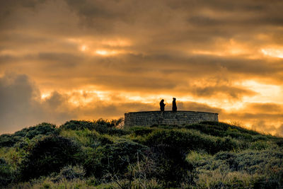 People standing on land against dramatic sky during sunset