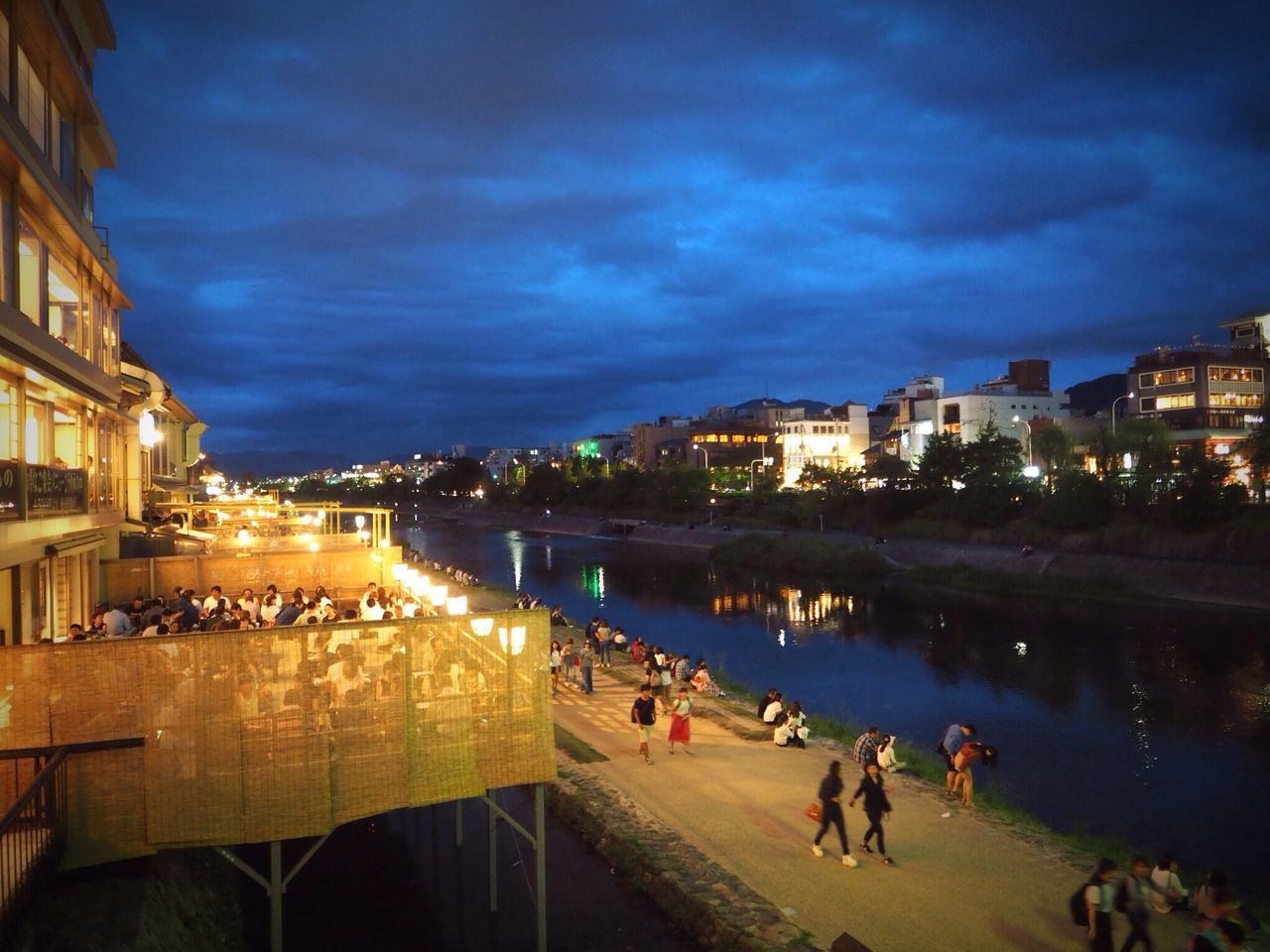 building exterior, illuminated, architecture, built structure, night, city, sky, large group of people, water, leisure activity, city life, river, person, cloud - sky, vacations, cloud, outdoors, blue, riverbank, town