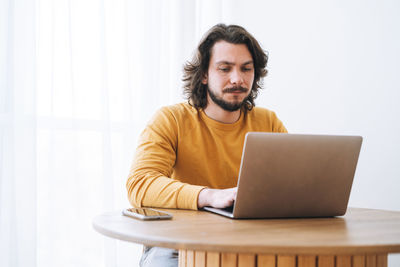 Young bearded brunette business man in casual yellow longsleeve working on laptop in modern office